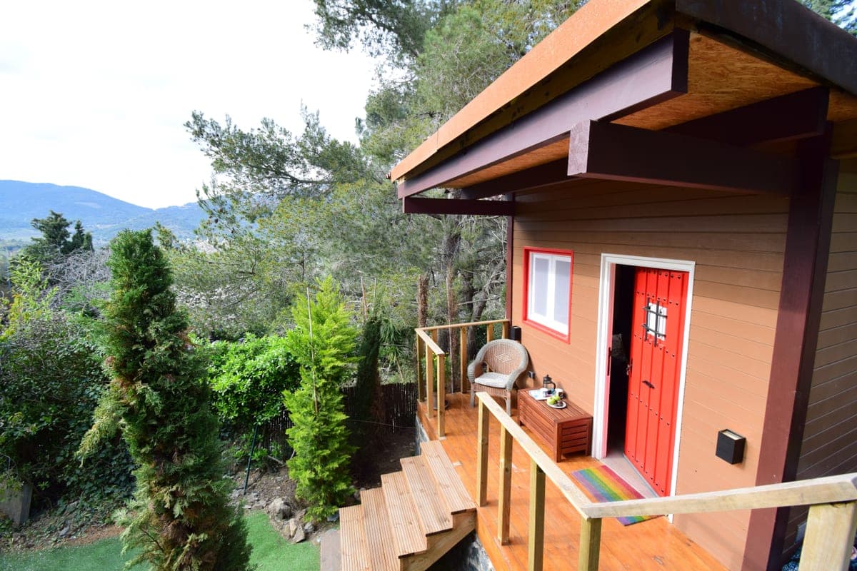 The guesthouse from the deck: the red door open, a chair and coffee pot, and the wooded valley behind.