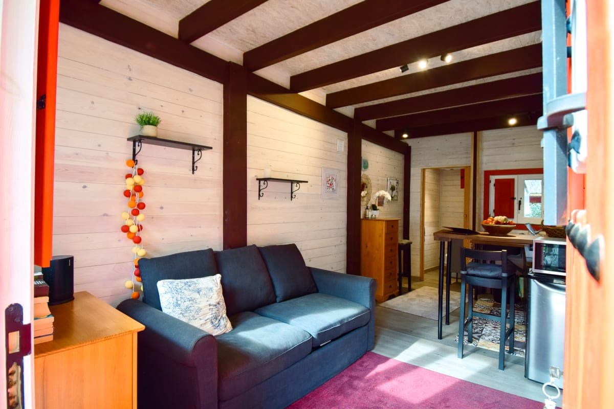 Inside the guesthouse: timber-lined walls under dark beams, the blue sofa bed, and the kitchenette with its breakfast bar beyond.