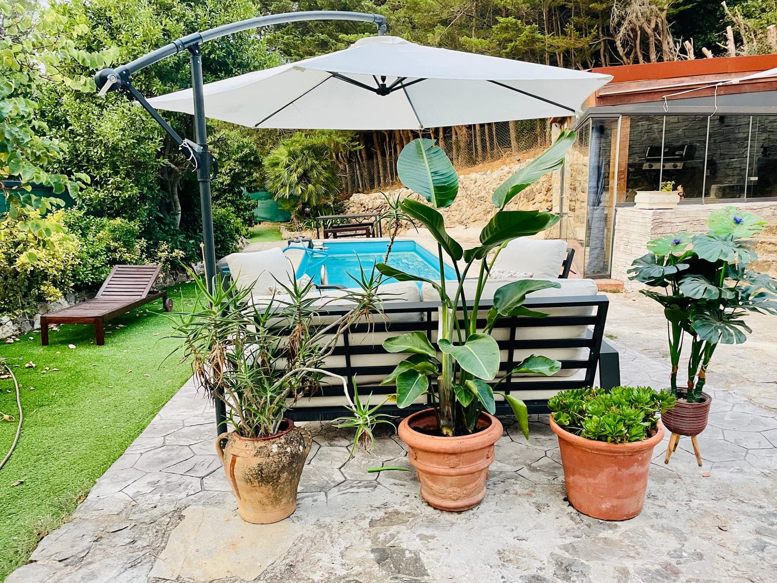 The pool terrace beside the stone house: the sofa set under a white parasol, potted plants, and the pool behind.