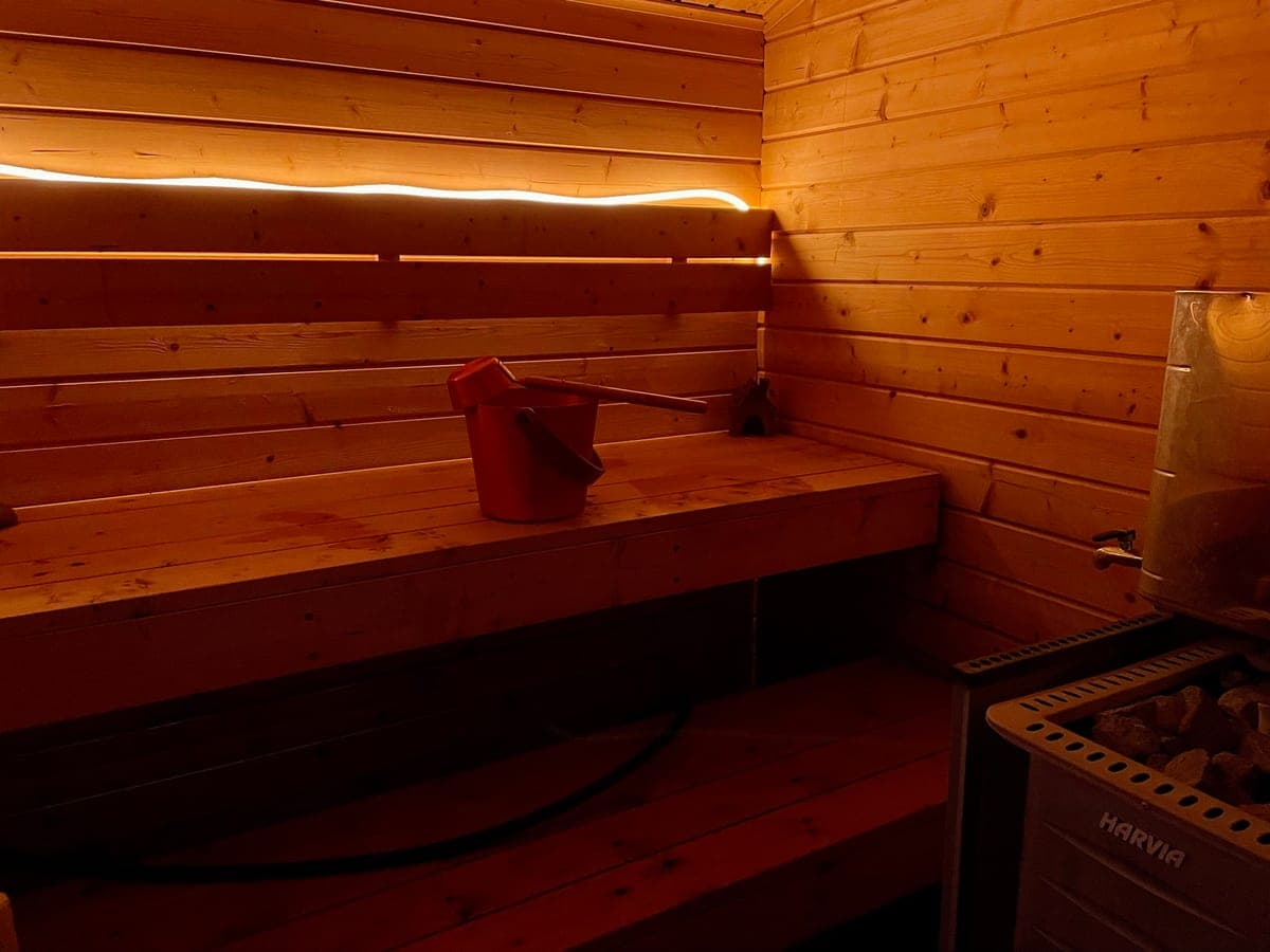 Inside the sauna: pine benches in low light, a red bucket and ladle, and the Harvia stove with its stones.