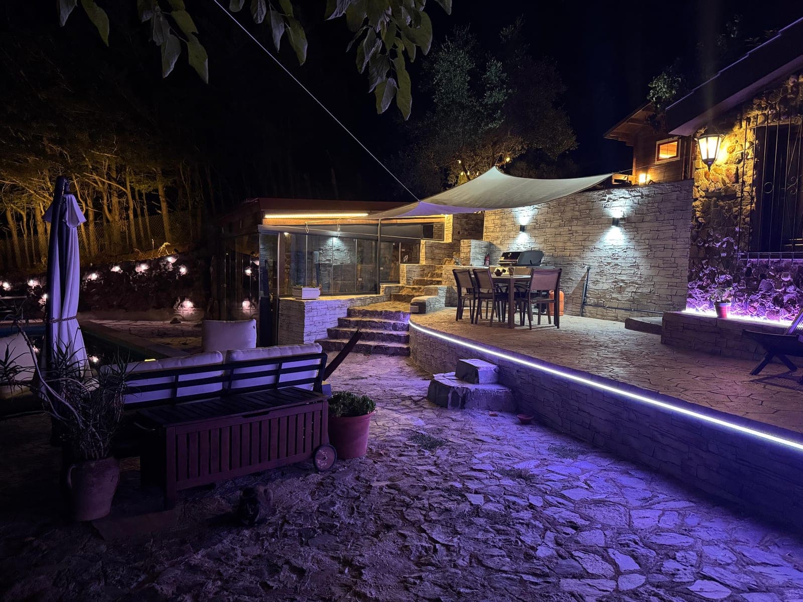 The terrace at night: the shade sail overhead, the stone wall, the dining table lit for the evening.