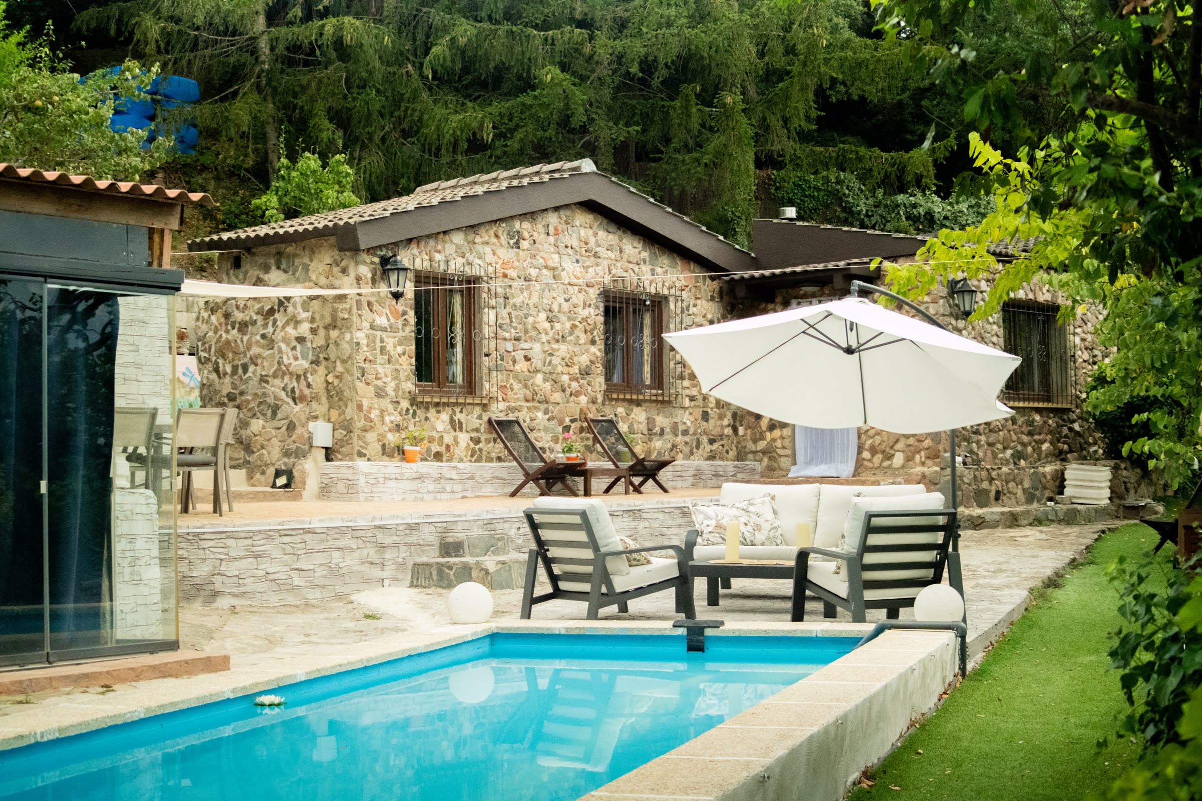 The stone house in daylight: its terrace and white parasol above the pool, and the wooded hillside behind.